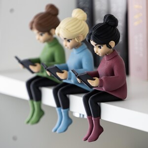 Könnte beinhalten: Drei Figuren von Frauen, die Bücher lesen, jede mit einer anderen Haarfarbe und einem anderen Pullover. Die Figuren sitzen auf einem weißen Regal, mit schwarzen Leggings und bunten Socken. Die Pullover sind grün, blau und weinrot.