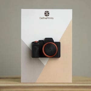 Op de afbeelding: Een zwart, camera-vormig object met een oranje lensring en bovenste knop is bevestigd aan een witte kaart met een geometrisch ontwerp. De kaart heeft de tekst "DefinePrints" bovenaan.