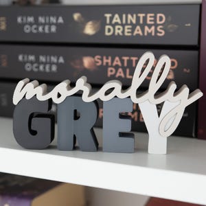 May include: A decorative wooden sign that spells out "morally GREY" in a mix of colors. The word "morally" is in a cursive font and the word "GREY" is in block letters. The sign is displayed on a white shelf with books in the background.