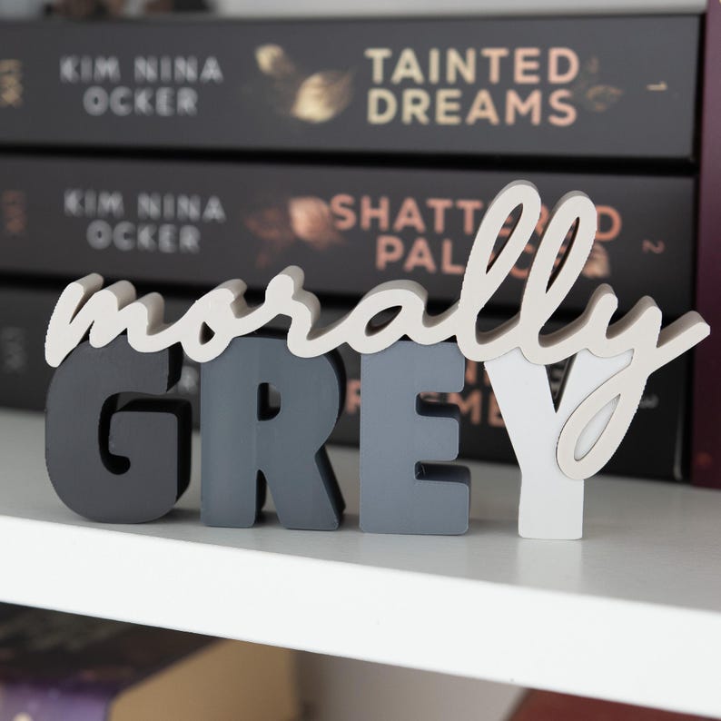 May include: A decorative wooden sign that spells out "morally GREY" in a mix of colors. The word "morally" is in a cursive font and the word "GREY" is in block letters. The sign is displayed on a white shelf with books in the background.