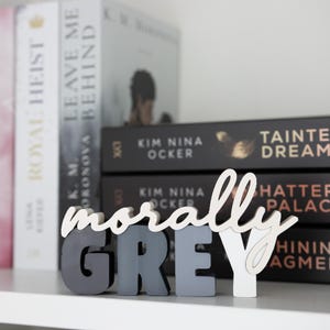 May include: A decorative wooden sign spelling "morally GREY" in a mix of gray and white. The sign is displayed on a shelf in front of a stack of books with titles like "Royal Heist" and "Leave Me Behind".