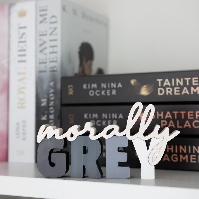 May include: A decorative wooden sign spelling "morally GREY" in a mix of gray and white. The sign is displayed on a shelf in front of a stack of books with titles like "Royal Heist" and "Leave Me Behind".