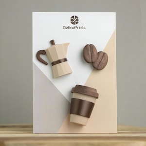 Op de afbeelding: Een display met decoratieve items met koffiethema. Inclusief een beige koffiezetapparaat, twee bruine koffiebonen en een koffiebeker voor onderweg met een bruine deksel en mouw. De tekst "DefinePrints" staat bovenaan.