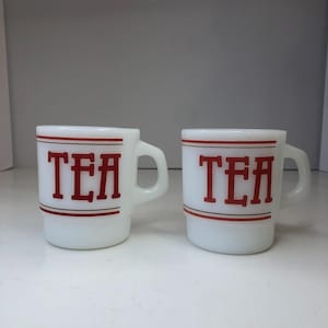 Könnte beinhalten: Zwei weiße Keramikbecher mit dem Wort "TEA" in roten Blockbuchstaben. Jeder Becher hat einen Henkel und einen roten Streifen oben und unten. Die Becher sind gestapelt und scheinen Vintage zu sein.