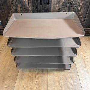 Könnte beinhalten: Ein Stapel von fünf Metalltabletts in verschiedenen Grautönen. Das oberste Tablett ist hellbeige, während die anderen dunkler grau sind. Die Tabletts haben eine rechteckige Form mit leicht erhöhten Kanten, was auf eine Organisation oder Servierfunktion hindeutet.