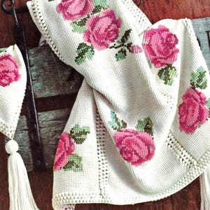 Könnte beinhalten: Cremefarbener, gehäkelter Stoff mit großen rosa Rosen und grünen Blättern. Das Design umfasst einen passenden Hut mit Quaste und dekorativem Rand. Die Rosen sind mit Kreuzstich gestickt, was dem Stoff Textur verleiht.