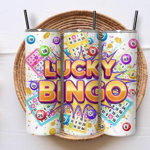 May include: Three white insulated tumblers with black straws, featuring a "Lucky Bingo" design. The design includes bingo cards, colorful bingo balls, and the text "Lucky Bingo" in a bold, yellow font. The tumblers are set on a woven basket.