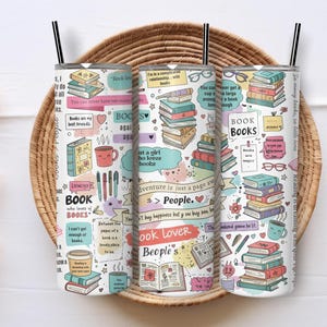 May include: Three white insulated tumblers with black straws, each adorned with colorful illustrations of books, coffee cups, pens, and text like "Book Lover" and "Adventure is just a page away." The tumblers are set in a woven basket.