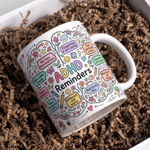 May include: A white ceramic mug featuring colorful doodle-style illustrations and text that reads ADHD Reminders, along with phrases like Not lazy. Overwhelmed., Forgetting not caring., Task paralysis is real., and Different still works.
