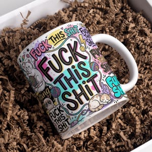 Könnte beinhalten: Ein weißer Keramikbecher mit einem farbenfrohen Grafikdruck und dem wiederholten Text "FUCK THIS SHIT" in fetter schwarzer Schrift. Das Design enthält Illustrationen von Katzen, Wollknäueln, Blitzen und Smileys in Pink, Blau und Gelb.