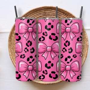 May include: Three pink tumblers with black straws, patterned with pink bows and black leopard spots. The tumblers are displayed on a woven basket.