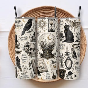May include: Three cylindrical tumblers with black straws, featuring a cream background and black illustrations of a raven, skull, crystal ball, hourglass, black cat, moth, snake, owl, mirror, and various occult symbols with text including Wild at Midnight.