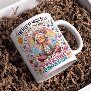 Puede incluir: Una taza de cerámica blanca con una ilustración colorida de una persona con cabello gris y gafas meditando en posición de loto. El texto dice THE PATH OF INNER PEACE BEGINS WITH FOUR WORDS y NOT MY F*CKING PROBLEM con ZERO F*CKS.