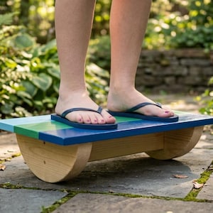 Puede incluir: Una tabla de equilibrio de madera rectangular con una superficie superior lisa pintada en franjas horizontales de color azul claro, verde y azul oscuro. La base consta de dos soportes de madera curvos en semicírculo.