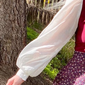 May include: A white long-sleeved blouse with a puffed sleeve and a frilled cuff. The blouse is made of a sheer fabric and is worn with a burgundy skirt with a floral print.