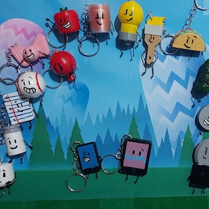 May include: A collection of colorful, handmade keychains featuring various cartoon-like characters. The keychains include a red apple, a yellow lightbulb, a taco, and a pickle, among others. Each keychain has a silver ring for attaching to keys or bags.