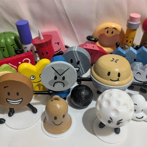 May include: A collection of small, hand-painted wooden figurines featuring various anthropomorphic characters with distinct facial expressions, including a golf ball, a bell, a coin, a pie, and a pencil, all standing on individual white circular bases.