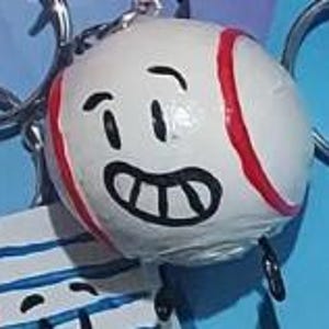 Puede incluir: Una pelota de béisbol blanca con costuras rojas y una cara de dibujos animados sonriente. La pelota tiene ojos negros y una sonrisa amplia y con dientes. Parece ser un artículo decorativo o un llavero.