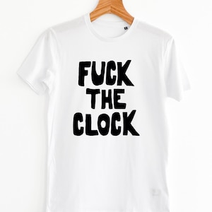 Puede incluir: Camiseta blanca con la frase "FUCK THE CLOCK" en letras negras y audaces. La camiseta está colgada en una percha de madera sobre un fondo blanco liso. El texto está centrado en la parte delantera.