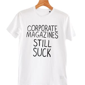 Puede incluir: Camiseta blanca con el texto negro "CORPORATE MAGAZINES STILL SUCK". La camiseta está colgada de una percha de madera. La camiseta es de cuello redondo.