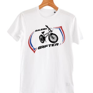 May include: A white short-sleeve t-shirt featuring a black graphic of a Raleigh Grifter bicycle. The design includes the text RALEIGH GRIFTER in black lettering, framed by two curved, brush-stroke style lines in red and blue on either side.