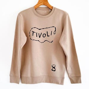 Puede incluir: Sudadera beige con la palabra "TIVOLI" en negro, delineada con una línea punteada. Debajo del texto hay un pequeño gráfico negro. La sudadera tiene mangas largas y cuello redondo.