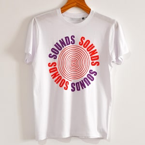 Peut inclure: T-shirt blanc avec un motif circulaire. Le mot "SOUNDS" est en rouge et violet, entourant une spirale rouge. Le t-shirt est présenté sur un cintre en bois.