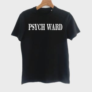 Könnte beinhalten: Schwarzes T-Shirt mit dem weißen Text "PSYCH WARD" auf der Brust. Der Text ist in einer fetten, Schablonen-artigen Schriftart gehalten. Das Shirt hängt an einem Holzkleiderbügel vor einem weißen Hintergrund.