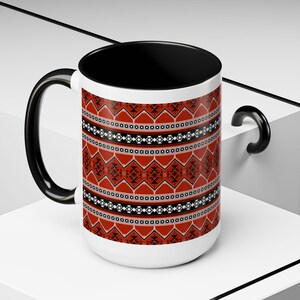 May include: A white ceramic mug with a black handle and interior. The mug features a repeating geometric pattern in red, black, and white. The design includes diamond and triangle shapes. The mug is on a white surface.