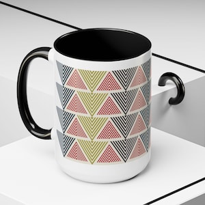 May include: White ceramic mug with a black interior and handle. The exterior features a repeating pattern of colorful triangles in red, green, and navy blue. The mug is displayed on a white surface.