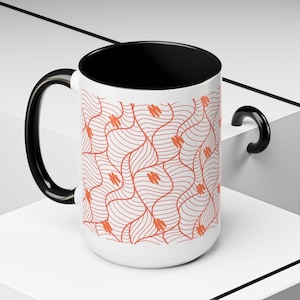 May include: A white ceramic mug with a black interior and handle. The exterior features a repeating pattern of wavy lines and small orange shapes. The mug is sitting on a white surface.