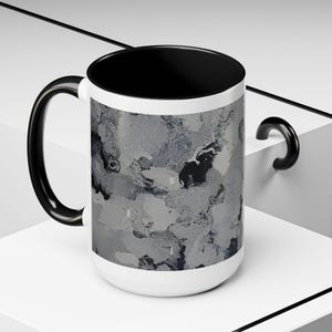 May include: White ceramic mug with a black interior and handle. The exterior features a gray and white abstract design. The mug is set against a white background with black accents.