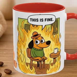 May include: White ceramic mug with a red interior and handle. The mug features a cartoon dog in a green sweater and hat, sitting in a burning room. A speech bubble says, "THIS IS FINE."