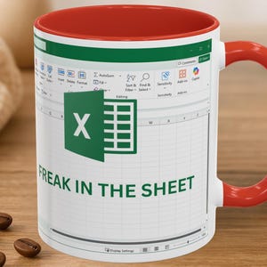May include: White ceramic mug with a red interior and handle. The mug features an Excel spreadsheet design with the text "FREAK IN THE SHEET" in green. Coffee beans are scattered on the wooden surface.