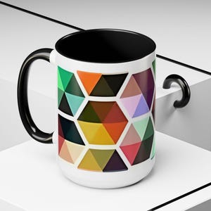 May include: White ceramic mug with a black interior and handle. The exterior features a geometric pattern of colorful triangles in shades of green, orange, yellow, pink, and purple. The mug is designed for hot or cold beverages.