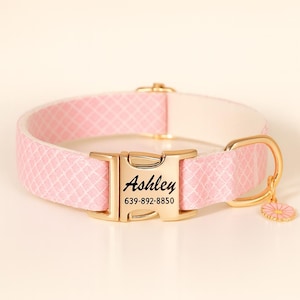 Puede incluir: Collar rosa para perro con hebilla y herrajes dorados. El collar presenta un patrón de diamantes y el nombre "Ashley" grabado en la hebilla, junto con un número de teléfono. Un pequeño dije de margarita cuelga del collar.