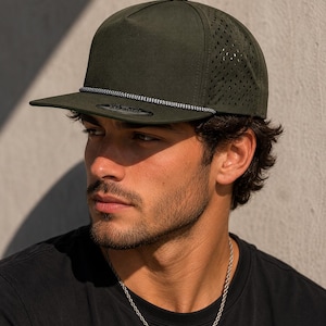 May include: An olive green five-panel snapback cap featuring a flat brim, a decorative braided cord across the base of the crown, and laser-cut perforated side panels for ventilation. A circular sticker is visible on the top of the flat brim.