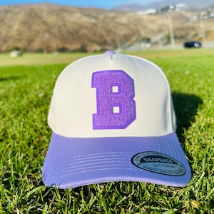 Puede incluir: Gorra snapback blanca y lila con una gran letra "B" lila bordada en la parte delantera. La gorra tiene una visera lila y está sobre hierba verde. La etiqueta dice "Snapback, Do it your way, Finest quality headwear."