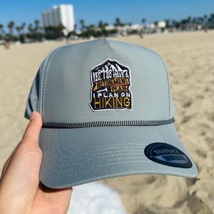 May include: Gray trucker hat with a black and white rope detail and a patch that reads "Yes, I Do Have A Retirement Plan, I Plan On Hiking." The hat has a curved brim and a snapback closure.