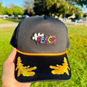 Puede incluir: Gorra de camionero negra con parte trasera de malla y visera curva. La gorra presenta un parche "PEACE" colorido y diseños bordados dorados en la visera. Un detalle de cuerda naranja acentúa la visera.