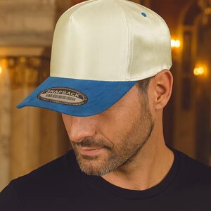 Puede incluir: Gorra snapback color crema con visera azul. La gorra tiene un acabado satinado y un botón azul en la parte superior. La visera lleva la etiqueta "SNAPBACK".