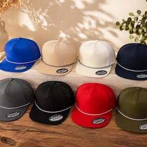 May include: A collection of eight snapback caps in various solid colors including blue, beige, white, navy, gray, black, red, and olive green. Each cap features a five-panel design, perforated side panels, a flat brim, and a decorative rope trim.
