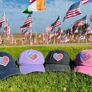 May include: Four baseball caps in navy, lavender, charcoal, and pink, each with a heart-shaped patch featuring the American flag. The caps are displayed on a grassy surface with numerous American flags waving in the background.