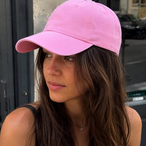May include: A solid light pink baseball cap with a structured crown, a curved brim, and stitched eyelets on the top panels. The cap features a classic six-panel design with a fabric-covered button at the center and a smooth, matte finish.