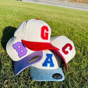 Puede incluir: Cuatro gorras de béisbol con viseras y letras de diferentes colores. Las gorras tienen coronas blancas y viseras de colores en rojo, morado y azul. Las letras son en rojo, morado, azul y rojo. Las gorras están sobre un lecho de hierba verde.