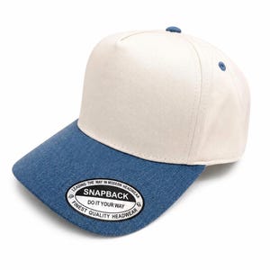Op de afbeelding: Een honkbalpet met vijf panelen, een crèmekleurige kroon en een blauwe klep met denimtextuur. Op de klep zit een ronde zwart-witte sticker met de tekst: LEADING THE WAY IN MODERN HEADWEAR SNAPBACK DO IT YOUR WAY FINEST QUALITY HEADWEAR.