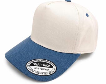 May include: A five-panel baseball cap with a cream-colored crown and a blue denim-textured brim. The brim features a circular black and white sticker that reads: LEADING THE WAY IN MODERN HEADWEAR SNAPBACK DO IT YOUR WAY FINEST QUALITY HEADWEAR.