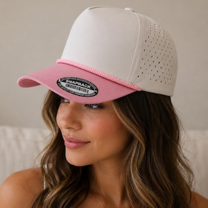 May include: A snapback cap featuring a solid off-white front panel, a pink brim, and a pink braided cord detail across the base of the crown. The side panels are off-white with a perforated design, and a black circular sticker is on the brim.