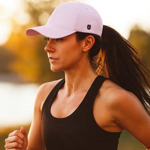May include: A light pink baseball cap with a curved brim and a small black logo on the side. The cap is worn by a person wearing a black tank top, running outdoors.