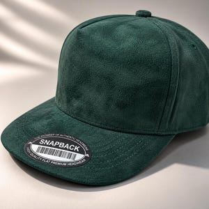 May include: A dark green suede-textured snapback cap with a five-panel construction and a flat brim. A circular black sticker is affixed to the brim, featuring the text "SNAPBACK" in white, along with a barcode and the words "THE PIONEER OF AUTHENTIC HEADWEAR" and "HIGH QUALITY FLAT PREMIUM HEADWEAR".